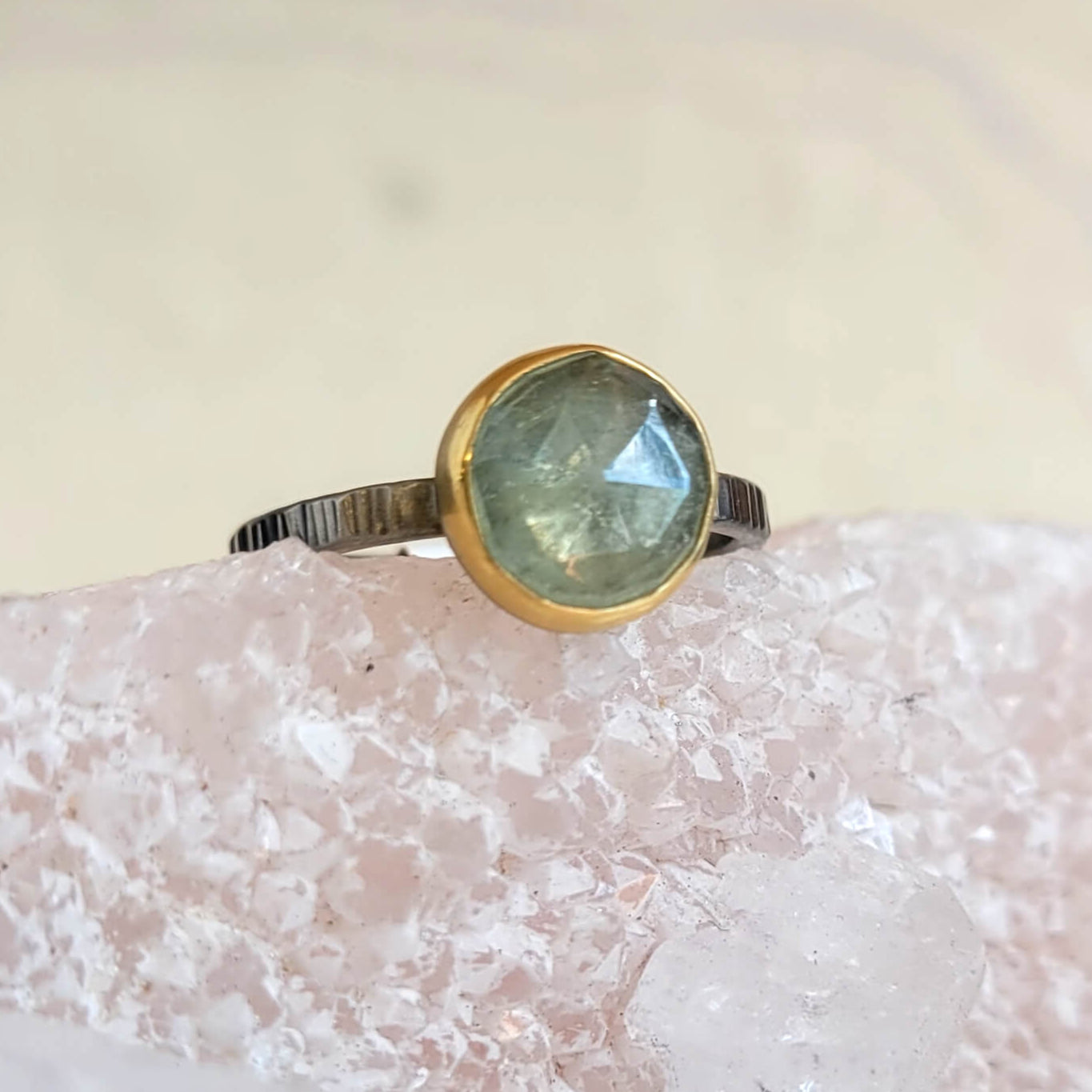 A pale green rose cut sapphire ring with a yellow gold bezel on a linear hammered palladium band, displayed on a crystal.