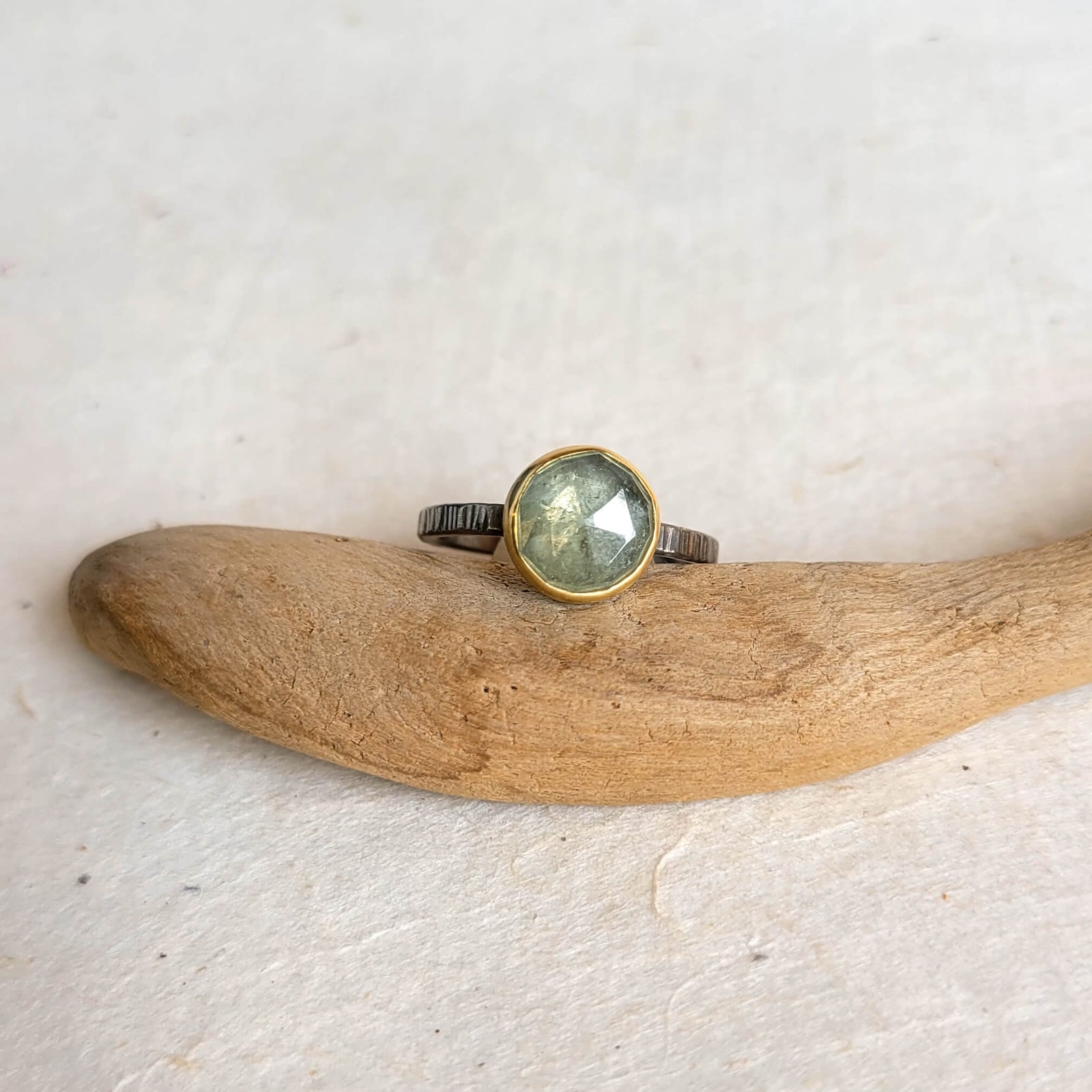 A pale green rose cut sapphire ring with a yellow gold bezel on a linear hammered palladium band, displayed on a crystal.