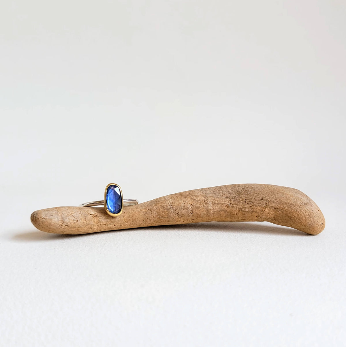 An irregular oval blue sapphire ring with a yellow gold and silver bezel set on a piece of driftwood.