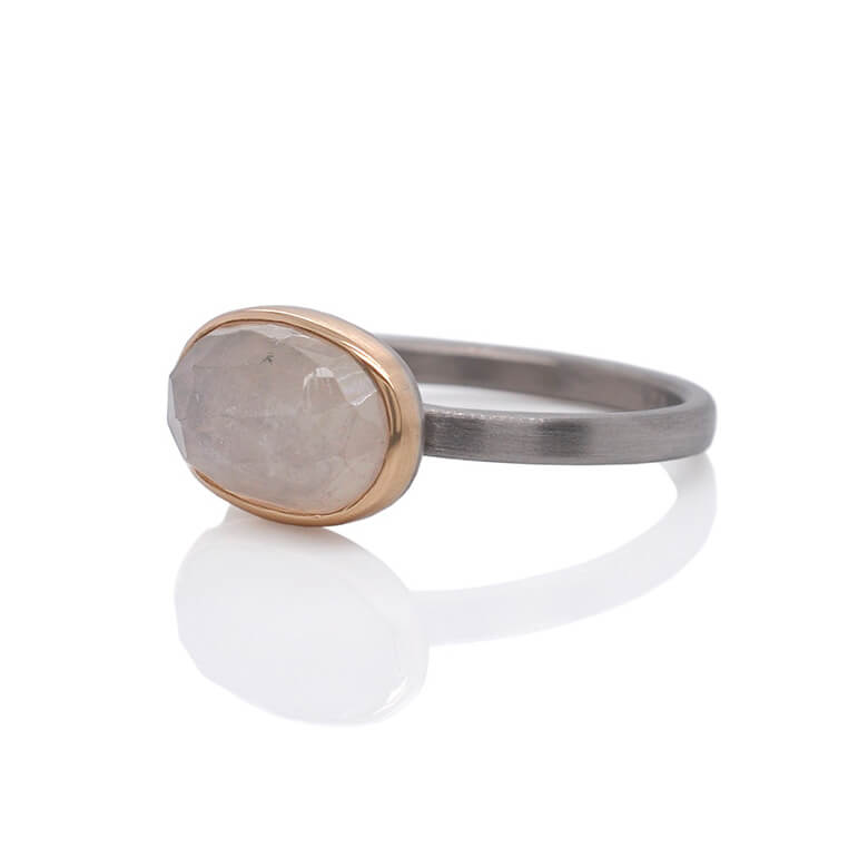 Cream colored oval sapphire ring with a yellow gold bezel on a palladium band.